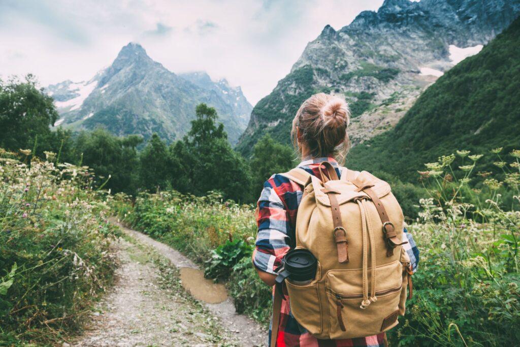 ine Wanderin mit einem großen Rucksack, die auf einem schmalen Pfad durch die Berge geht. Sie genießt die unberührte Natur und die Aussicht auf die hohen Berggipfel.