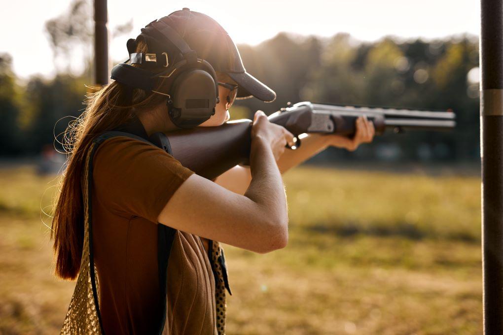 Eine Frau beim Schießtraining im Freien, sie hält ein Gewehr und trägt Gehörschutz. Sie fokussiert sich auf ihr Ziel.