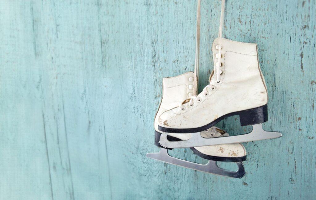 Weisse klassische Eiskunstlauf-Schuhe haengen vor blauer Holzbrettwand – nostalgische Symbolik fuer moderne Bauer Schlittschuhe
